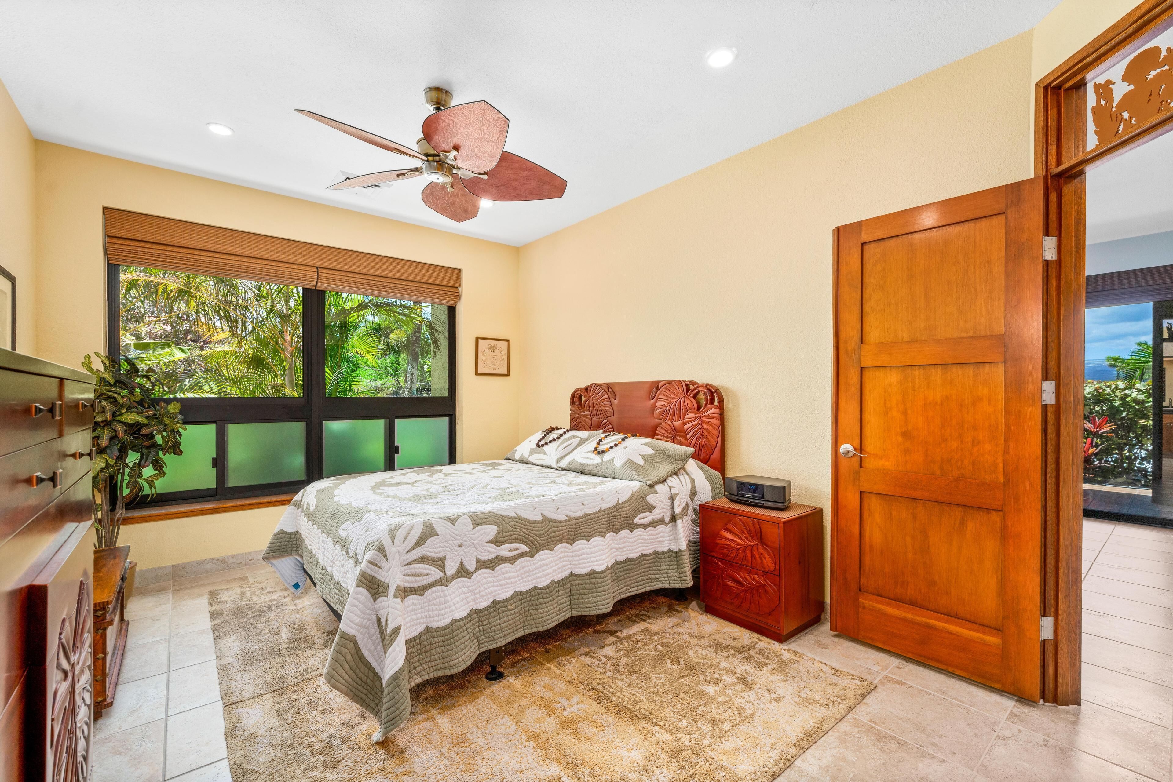 A bedroom with a bed, a nightstand, a wooden door, a ceiling fan, and a window with a view of greenery.