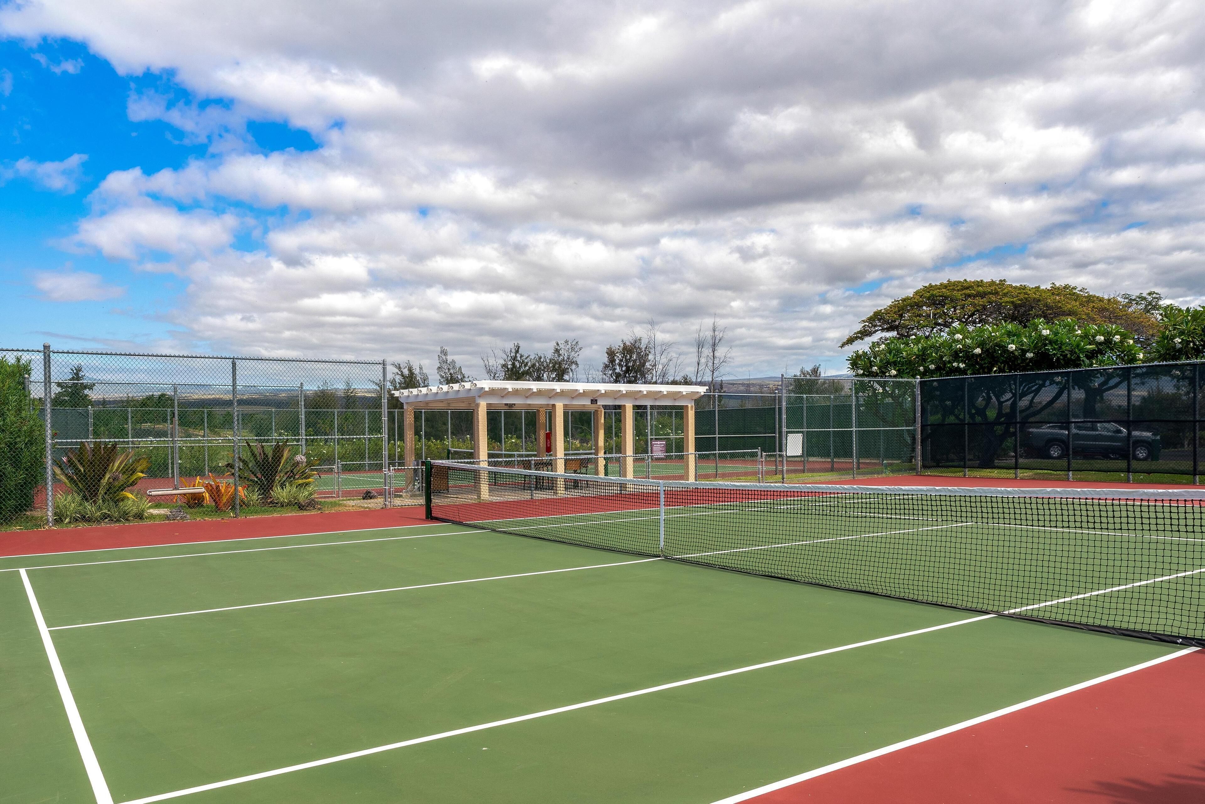 Un court de tennis avec un filet, entouré d’une clôture, et une zone de sièges abritée.