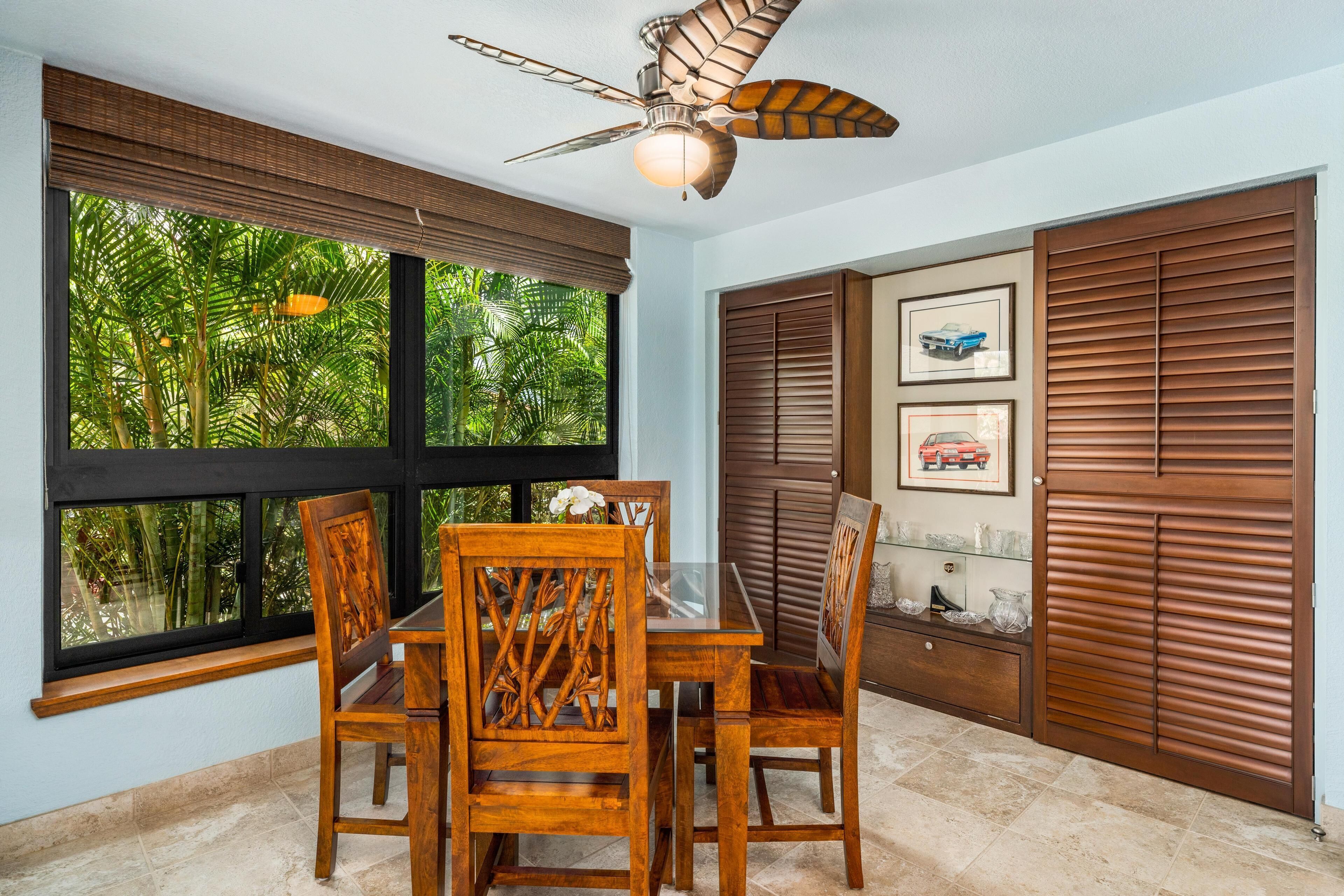 A dining area with a table and chairs, a ceiling fan, and a view of lush greenery through large windows.