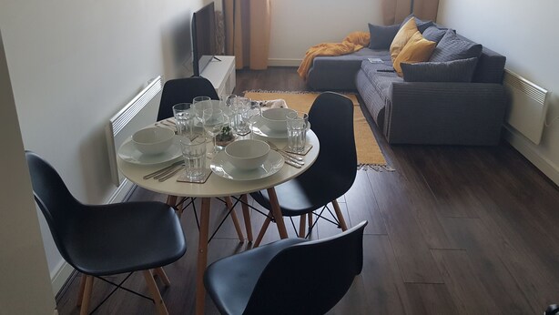 Een eethoek met een ronde tafel voor vier personen, omringd door zwarte stoelen. Op de achtergrond staat een grijze bank met een gele plaid.