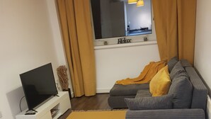 Een woonkamer met een bank, een televisie op een dressoir, een geel vloerkleed en een houten vloer.