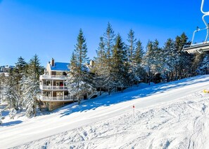 Une piste de ski enneigée, avec un immeuble à plusieurs étages doté de balcons, le tout entouré d’arbres chargés de neige.