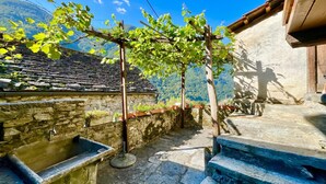 Un patio con paredes de piedra, un lavamanos de piedra, escalones de madera y una pérgola cubierta de plantas trepadoras.