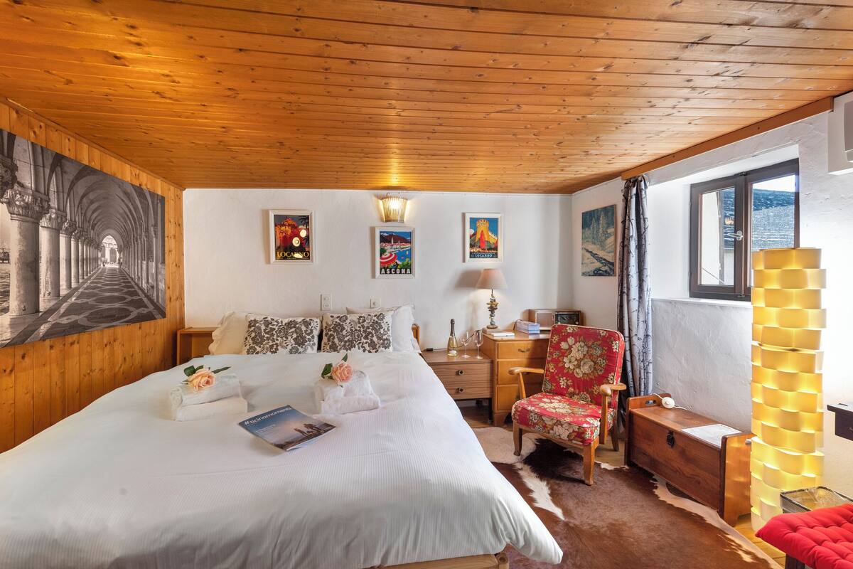 A bedroom with a wooden ceiling, a large bed with white bedding, a wooden nightstand, a chair, a lamp, and framed pictures on the wall.