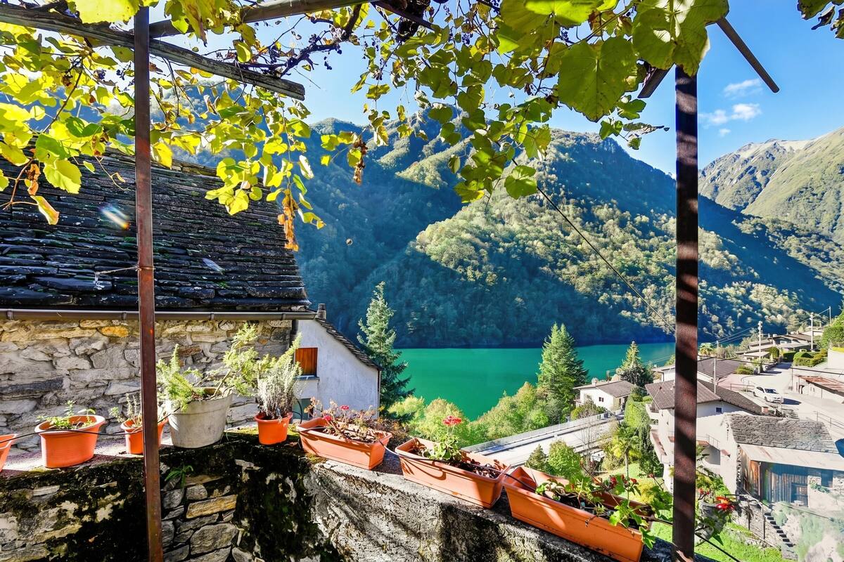 A stone building with terraced potted plants, a clear view of a lake, and surrounding mountains.