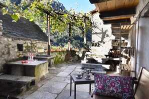 A rustic outdoor seating area with stone tables and benches, surrounded by stone walls and a wooden pergola.