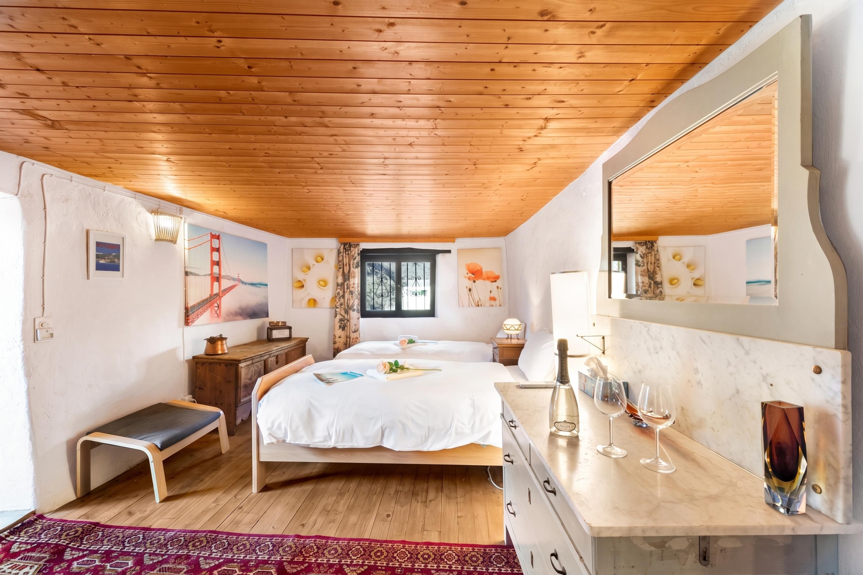 Un dormitorio con techo de madera, una cama con ropa de cama blanca, un aparador, un espejo y una ventana con cortinas.