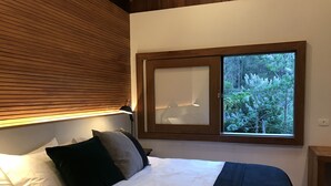 A bedroom with a bed, pillows, a nightstand, a lamp, and a window with a view of greenery. - Chalés Realli (Bocaina de Minas)