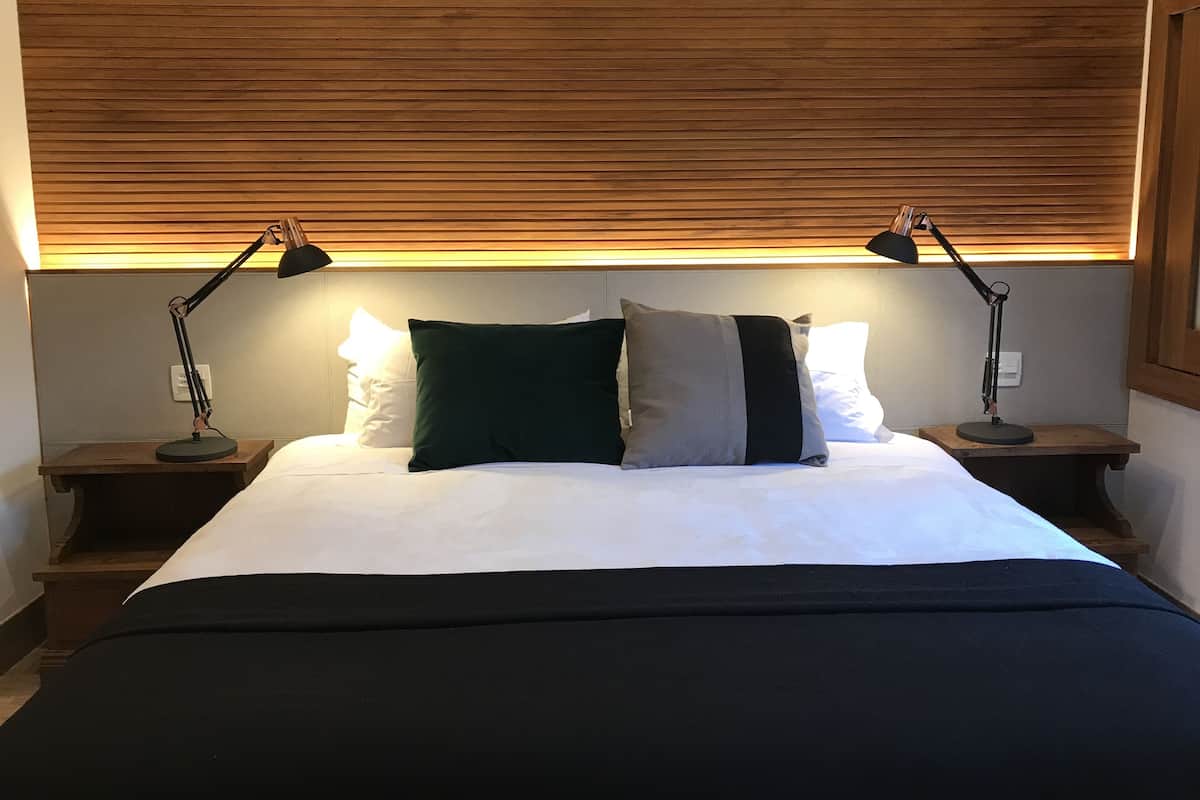 A neatly made bed with a white duvet and dark green and grey pillows, flanked by two bedside tables with lamps, in a room with wooden paneling on the ceiling.