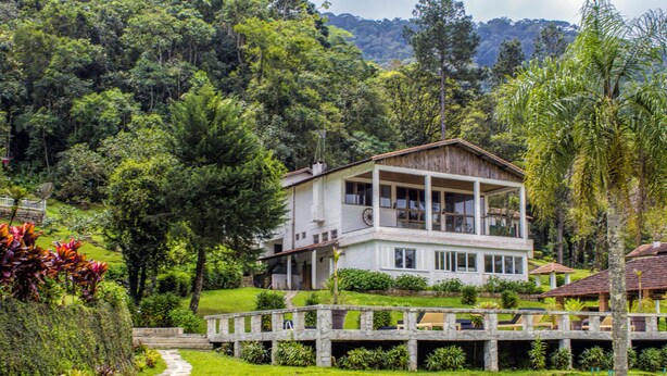 A two-story house with a balcony, surrounded by lush greenery and a stone bridge. - Chalés Realli (Bocaina de Minas)