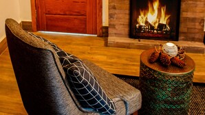 A room with a wooden door, a brick fireplace with a fire, a grey upholstered chair with a patterned pillow, and a round table with pine cones and a candle. - Chalés Realli (Bocaina de Minas)