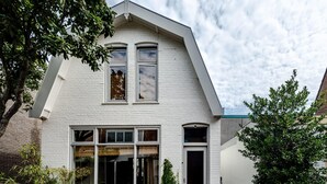 Ein weißes, zweistöckiges Haus mit Satteldach, großen Fenstern und einer Veranda mit Sitzgelegenheit.