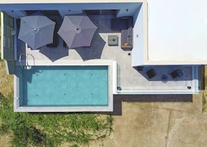 Vista aérea de uma área de piscina moderna ao ar livre, com uma piscina retangular, dois guarda-sóis e um espaço de estar com almofadas.