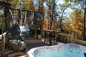 A wooden deck with a hot tub, outdoor seating, and a swing chair overlooking a forest.