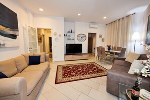 Uma sala de estar com um sofá bege, um sofá marrom, uma televisão e um espaço de jantar com cadeiras e uma mesa.
