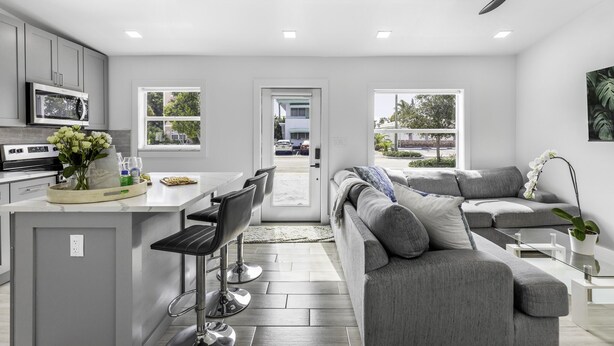 A modern kitchen with grey cabinets, a central island with bar stools, and a grey sofa in the living area.