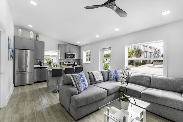 A modern kitchen with stainless steel appliances, a grey sofa with patterned cushions, and a glass coffee table.