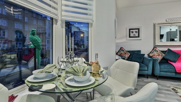 A modern living room with a glass dining table set for four, a green parrot sculpture, a blue sofa with colorful cushions, and a city view through large windows.
