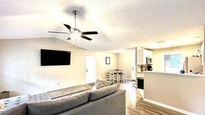Un moderno salón con un sofá modular gris, un ventilador de techo, un televisor de pantalla plana montado en la pared y una zona de cocina con microondas y refrigerador.