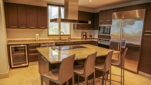 A modern kitchen with dark cabinetry, a large island with bar stools, stainless steel appliances, and a window providing natural light. - Waking up in a tropical reserve with the sound of the sea caressing your soul. (Río Grande)