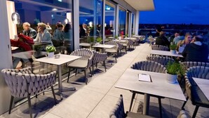 Un área de comedor al aire libre con mesas y sillas, ventanas amplias y vistas a la ciudad.