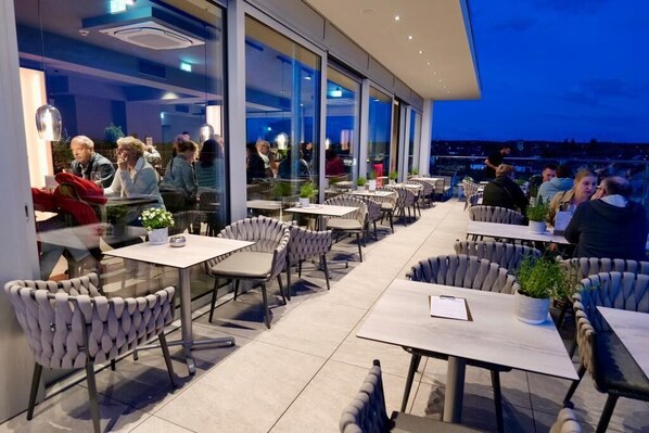 An outdoor dining area with tables and chairs, large windows, and a view of the cityscape. - Hotel Kump 365 (Paderborn)