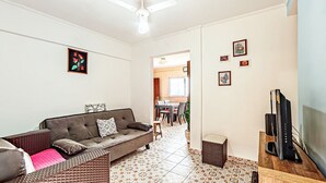 A living room with a sofa, a television, and a dining area with a table and chairs. - Agradável apartamento no Caiçara (Praia Grande)