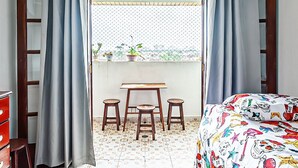 A bedroom with a bed, a desk, two chairs, a ceiling fan, and a view through a window with curtains. - Agradável apartamento no Caiçara (Praia Grande)