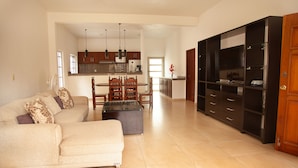 A spacious living room with a beige sofa, a coffee table, and a dining area with a wooden table and chairs. There is a kitchen area with cabinets and a refrigerator in the background. - CASA BENCHITO (Tulum)