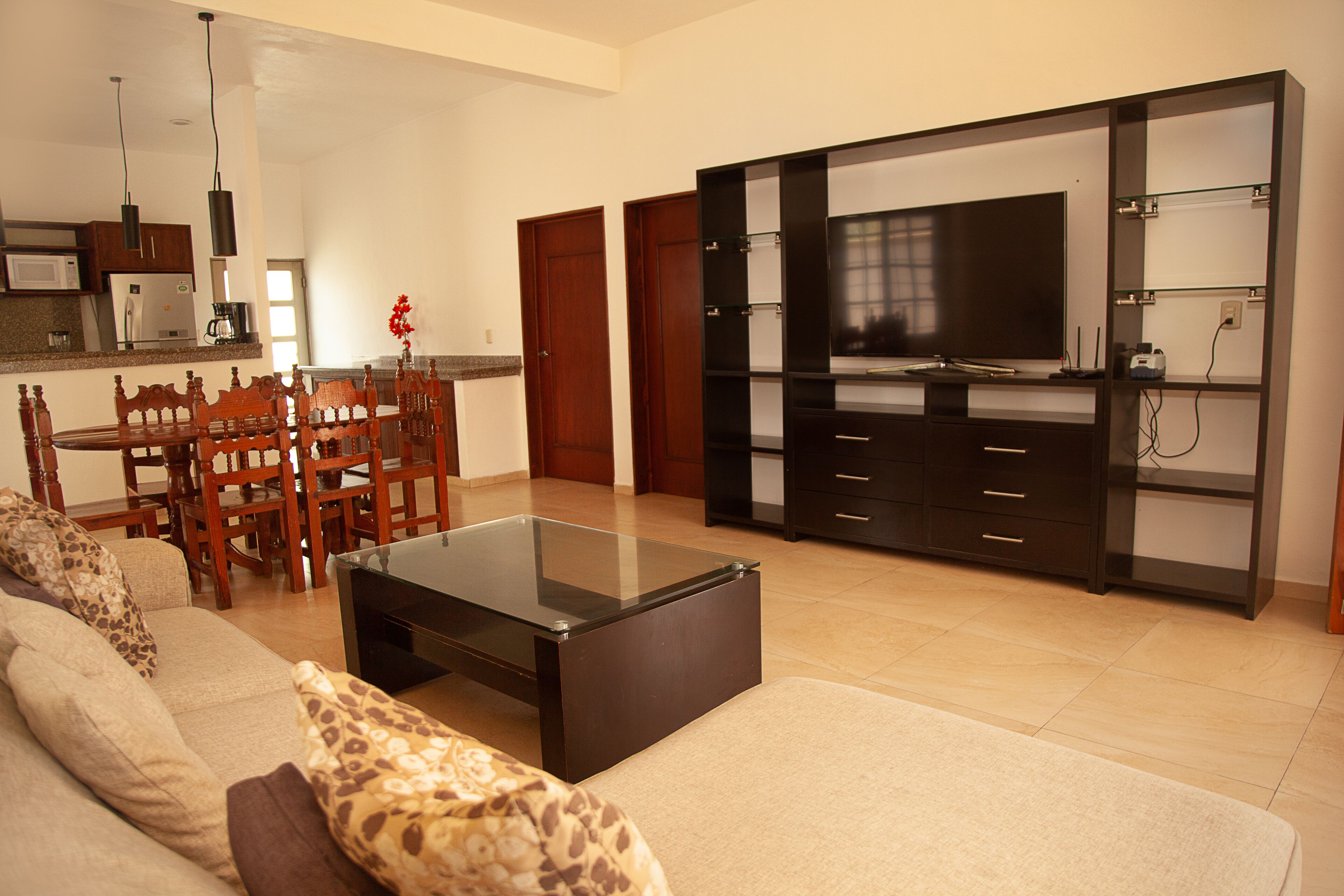 A living room with a sofa, a glass coffee table, a dining area with chairs, a kitchen with appliances, and a TV on a cabinet.