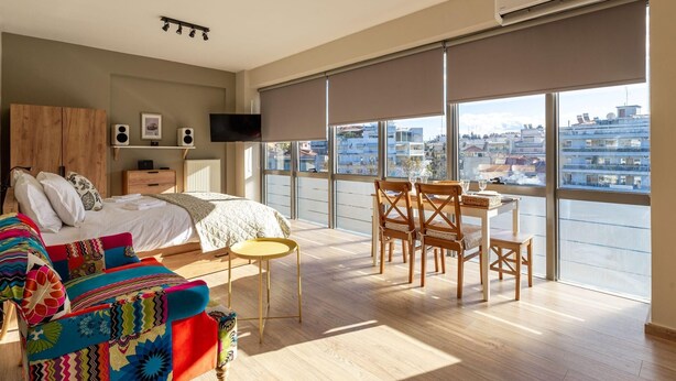Une chambre moderne avec une grande fenêtre, un lit, un canapé coloré et un coin repas avec des chaises.