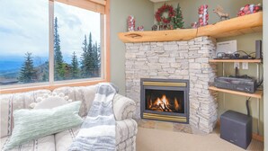 A cozy living room with a stone fireplace, a large window overlooking a forest, and a fireplace mantel with holiday decorations.