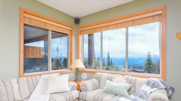 A cozy living room with a sectional sofa, large windows with wooden frames, and a view of mountains and trees.