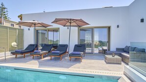 Un espace piscine extérieur moderne, doté de chaises longues et de parasols, d’un espace détente fermé par des baies vitrées et offrant une vue dégagée sur le paysage environnant.