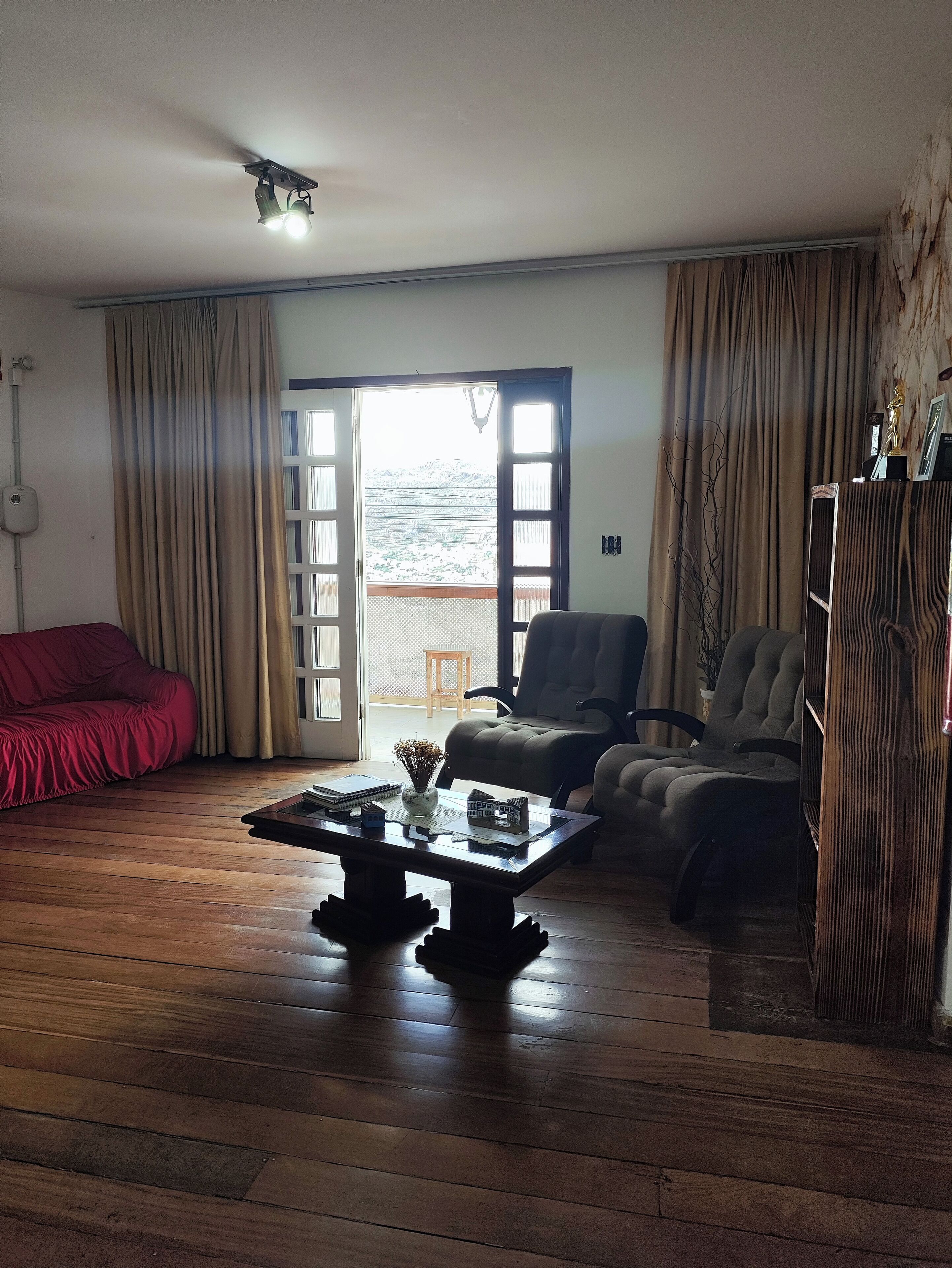 Uma sala de estar com piso de madeira, um sofá vermelho, uma poltrona verde, uma mesa de centro de madeira com casas de brinquedo e vista para uma varanda através de uma porta de vidro.