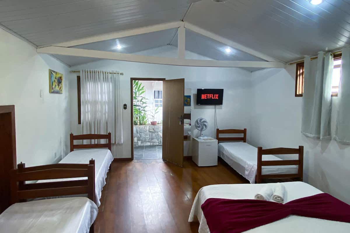 A room with four single beds, a ceiling fan, a television displaying 'NETFLIX', and a small table with a fan.