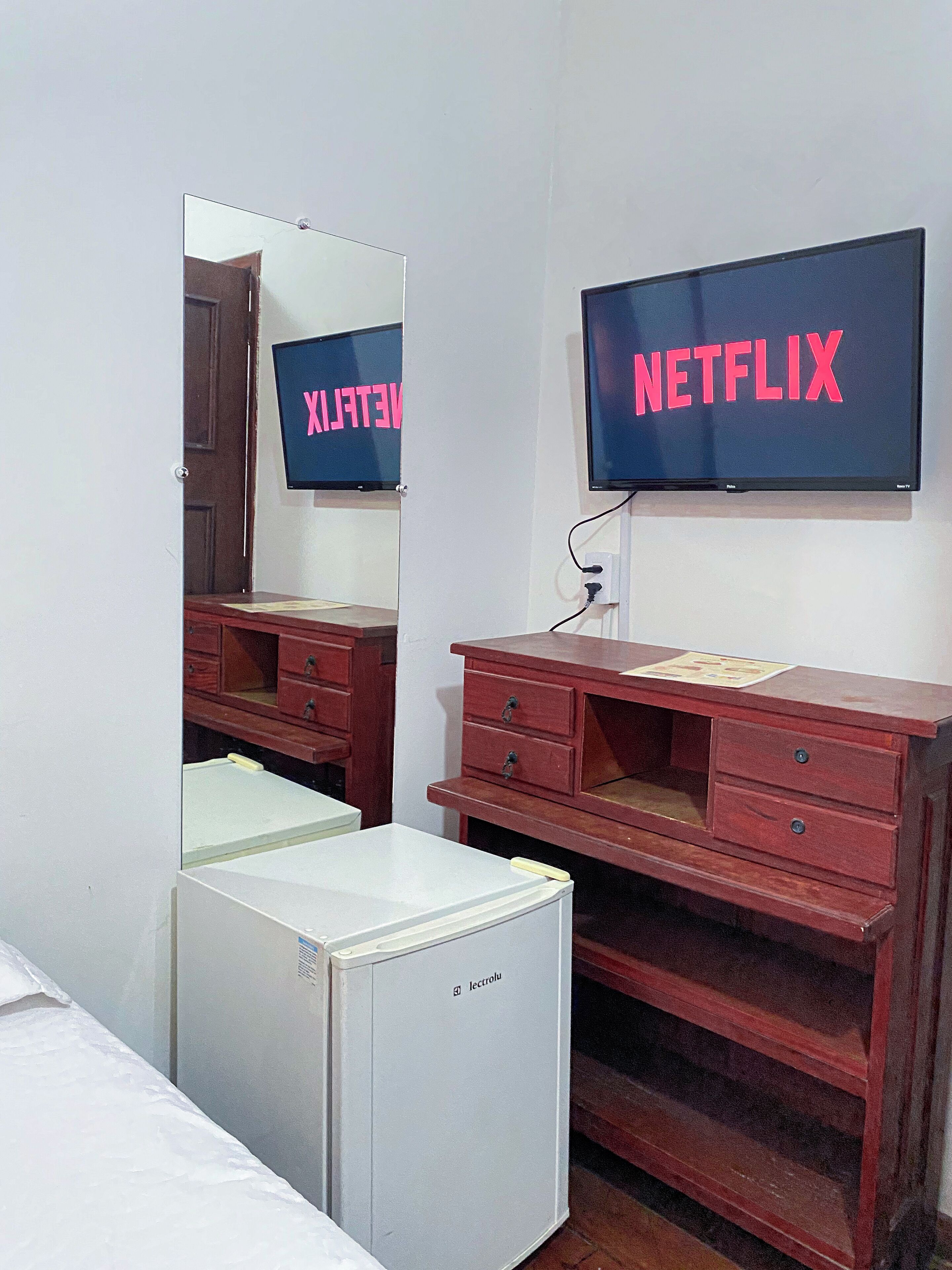 Una habitación con un refrigerador blanco, un escritorio de madera con cajones, un espejo y un televisor montado en la pared que muestra el logotipo de Netflix.