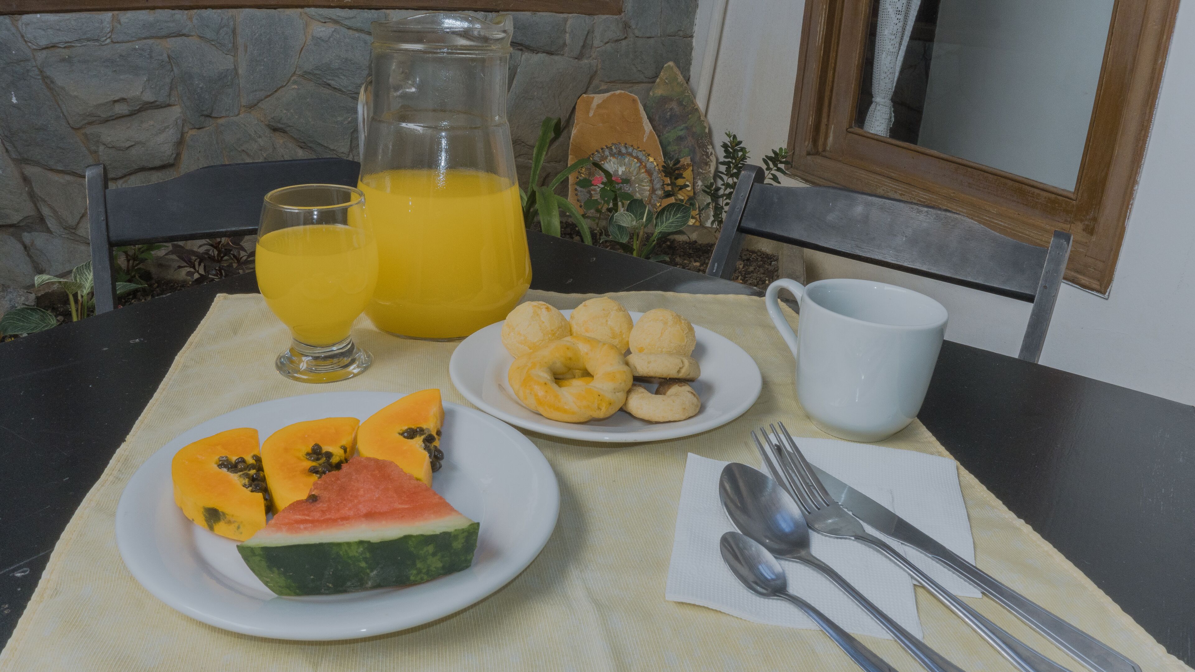 A breakfast setting with a stone wall, a table with a placemat, plates of fruit, pastries, a glass of orange juice, a mug, cutlery, and a pitcher.