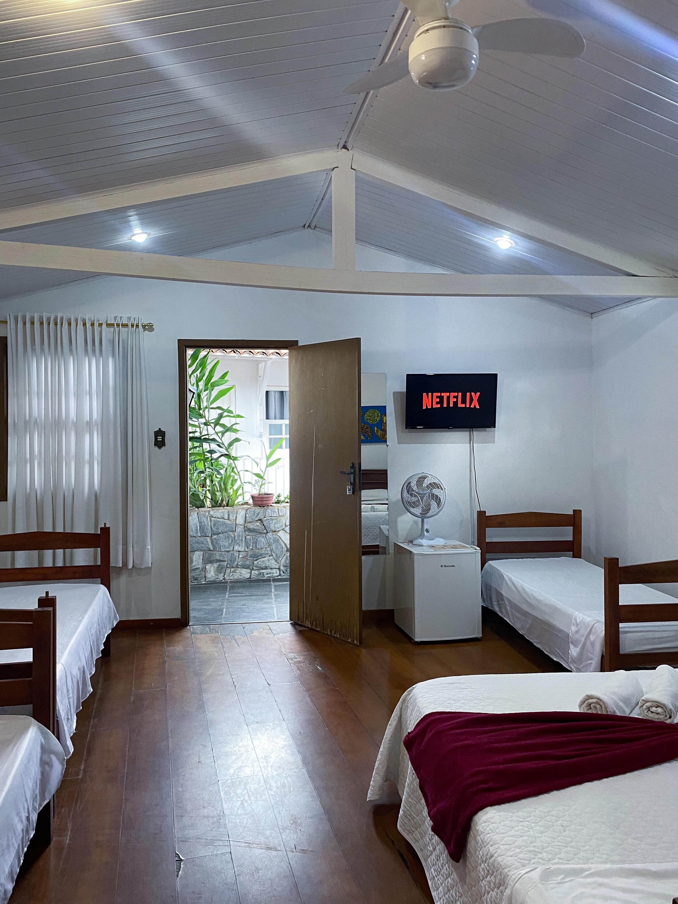 Habitación de hotel con dos camas, un televisor mostrando 'NETFLIX', un ventilador pequeño y vistas de un jardín a través de una puerta abierta.