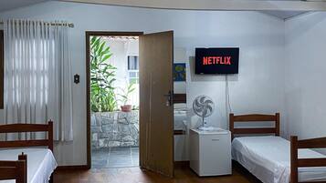 A hotel room with two beds, a television displaying 'NETFLIX,' a small fan, and a view of a garden through an open door.