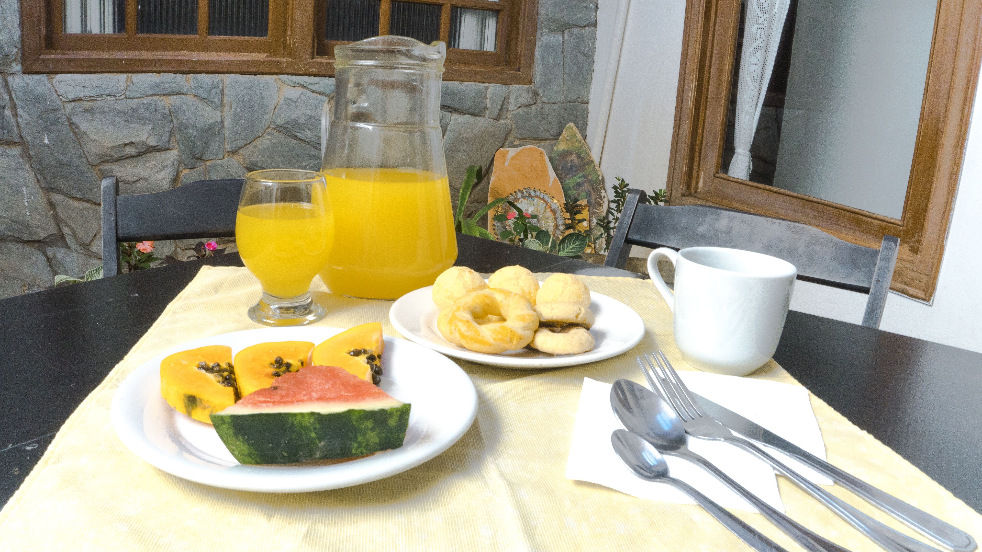 Una mesa de desayuno con una fuente de sandía en rodajas, una jarras de jugo de naranja, un vaso de jugo de naranja y una fuente de cuajada.