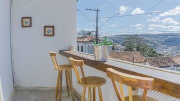Un balcón con una barra de madera y dos sillas de madera, un cubo metálico con botellas, y cuadros en la pared.