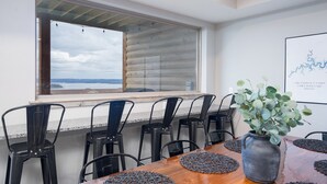 Un comedor con una mesa de madera, sillas negras, un jarrón con plantas verdes, una ventana con vista al mar y una obra de arte enmarcada en la pared.