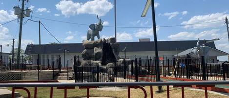 Uma área cercada com uma grande escultura de elefante, um edifício ao fundo e um céu azul claro.