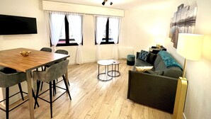 Een moderne woonkamer met een houten eettafel, grijze stoelen, een grijze bank en een aan de muur bevestigde televisie.