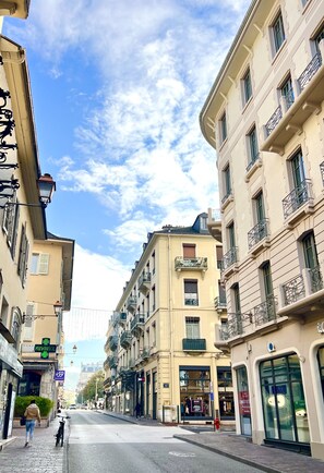 Une rue bordée de bâtiments des deux côtés, un ciel dégagé et une personne qui marche en poussant un vélo.
