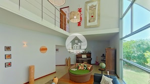 A spacious living room with a high ceiling, a large window, and a view of greenery outside. There is a wooden coffee table with books and decorative items, a blue sofa with cushions, and a wicker chair.