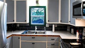 Une cuisine avec des meubles gris, un réfrigérateur en acier inoxydable, un four à micro-ondes et une grande fenêtre donnant sur les arbres.