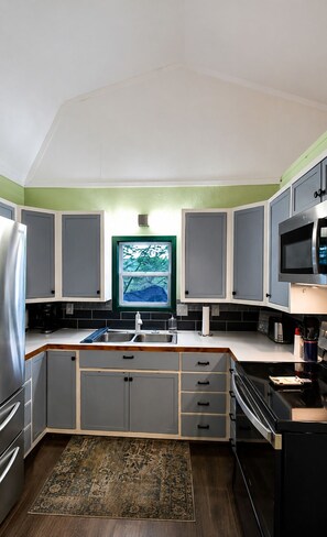 A kitchen with grey cabinets, a stainless steel refrigerator, a microwave, and a large window with a view of trees.