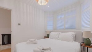 A hotel room with a large bed, a bedside table, a lamp, and a window with blinds. - Stylish Garden Home 2 BDR Wimbledon (London)